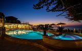 Stimmungsvolle Abendkulisse am beleuchteten Pool mit Whirlpool, gemütlichen Sitzplätzen und Blick auf den farbenprächtigen Sonnenuntergang über dem Meer.