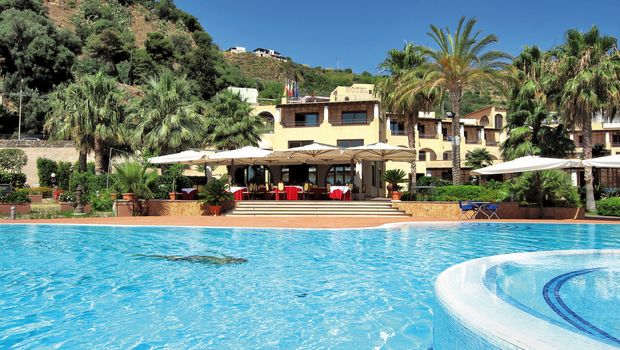 Großer Außenpool vor einem mediterranen Hotelgebäude mit Sonnenterrasse, Palmen und grüner Hügellandschaft im Hintergrund – sonnige Urlaubsatmosphäre unter blauem Himmel.