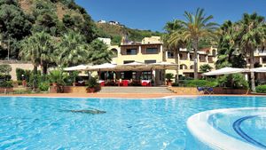 Großer Außenpool vor einem mediterranen Hotelgebäude mit Sonnenterrasse, Palmen und grüner Hügellandschaft im Hintergrund – sonnige Urlaubsatmosphäre unter blauem Himmel.