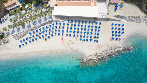 **Alt-Text:** Luftaufnahme eines Strandes mit türkisblauem Wasser und vielen blauen Sonnenschirmen.