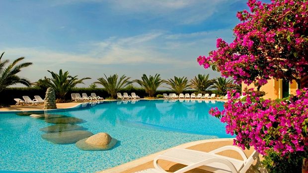 Runder Pool mit Natursteininsel, gesäumt von Palmen und weißen Liegestühlen – im Vordergrund ein blühender Bougainvillea-Strauch.