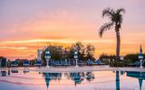 Poollandschaft mit blauen Sonnenliegen und Palmen vor einem farbenprächtigen Sonnenuntergang mit Blick auf das Meer und eine entfernte Insel am Horizont.