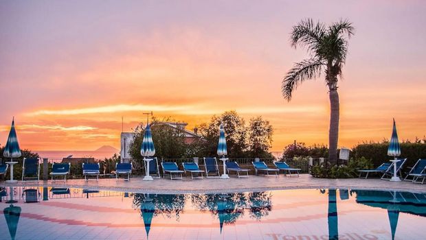 Poollandschaft mit blauen Sonnenliegen und Palmen vor einem farbenprächtigen Sonnenuntergang mit Blick auf das Meer und eine entfernte Insel am Horizont.