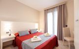 Modern eingerichtetes Doppelzimmer mit rotem Bettüberwurf, grauen Dekokissen und Zugang zum Balkon durch eine bodentiefe Tür mit Vorhängen.