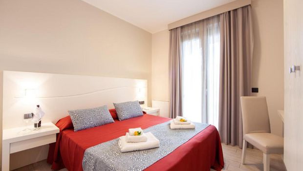 Modern eingerichtetes Doppelzimmer mit rotem Bettüberwurf, grauen Dekokissen und Zugang zum Balkon durch eine bodentiefe Tür mit Vorhängen.