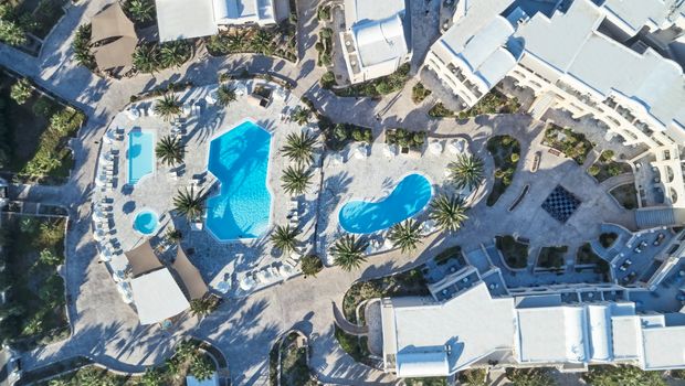 Luftaufnahme einer weitläufigen Hotelanlage mit mehreren Pools, Palmen, Liegeflächen und hellen Gebäuden im kykladischen Stil – harmonisch eingebettet in eine mediterrane Landschaft.