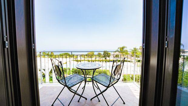 Balkon mit zwei Eisenstühlen und Tisch, Blick auf Poolanlage und das Meer in der Ferne.
