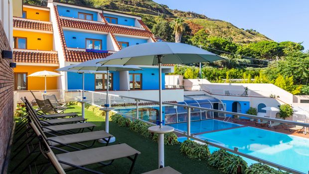 Moderne Hotelanlage mit bunten Fassaden, sonniger Liegewiese und großzügigem Poolbereich inmitten grüner Hügellandschaft.