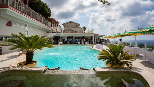Einladender Poolbereich mit Blick aufs Meer – Palmen und Sonnenschirme schaffen ein entspanntes Urlaubsambiente, während Gäste sich im Wasser oder auf der Terrasse amüsieren.