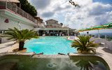 Einladender Poolbereich mit Blick aufs Meer – Palmen und Sonnenschirme schaffen ein entspanntes Urlaubsambiente, während Gäste sich im Wasser oder auf der Terrasse amüsieren.