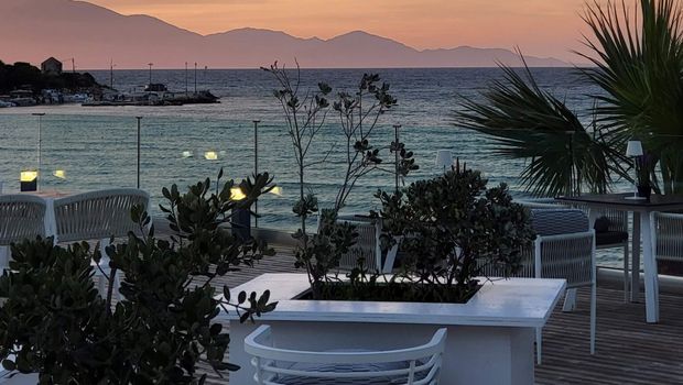 Stimmungsvolle Terrasse mit modernen Sitzmöbeln und Palmen, direkt am Meer gelegen – im Hintergrund ein farbenprächtiger Sonnenuntergang über den Bergen.