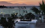 Stimmungsvolle Terrasse mit modernen Sitzmöbeln und Palmen, direkt am Meer gelegen – im Hintergrund ein farbenprächtiger Sonnenuntergang über den Bergen.