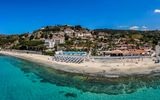 Panoramablick auf eine Hotelanlage direkt am klaren, türkisblauen Meer mit Liegestühlen am Sandstrand.