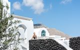 Weiße Hotelarchitektur im kykladischen Stil mit Natursteinmauern und Bergkulisse im Hintergrund – eine Frau genießt die Sonne vor strahlend blauem Himmel.
