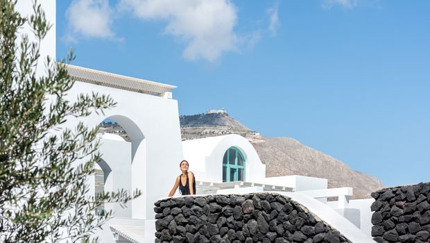 Weiße Hotelarchitektur im kykladischen Stil mit Natursteinmauern und Bergkulisse im Hintergrund – eine Frau genießt die Sonne vor strahlend blauem Himmel.