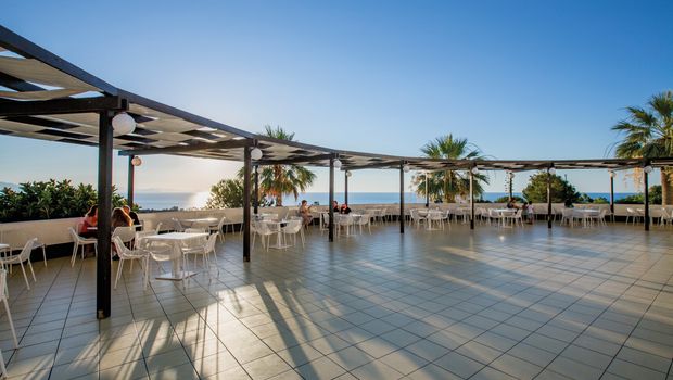 Großzügige Terrasse mit weißen Tischen und Stühlen unter Pergolen – ideal zum Speisen oder Entspannen mit Panoramablick auf das Meer und die untergehende Sonne.