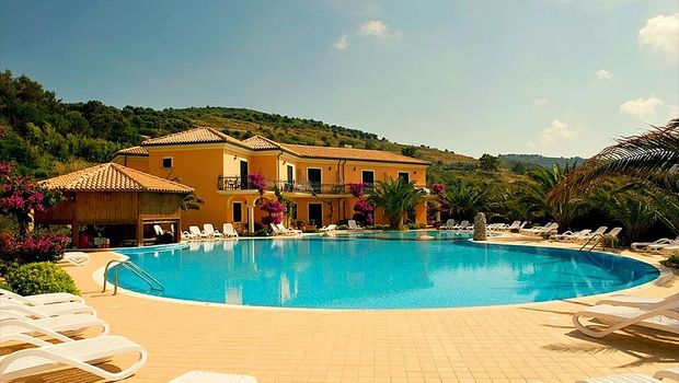 Großzügiger Poolbereich mit zahlreichen Liegestühlen, umgeben von Palmen und mediterraner Vegetation vor einer gelb gestrichenen Hotelanlage.
